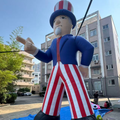 A large inflatable Uncle Sam-style patriotic figure stands outdoors in front of apartment buildings, wearing a red, white, and blue outfit with a striped top hat and pointing with one hand.